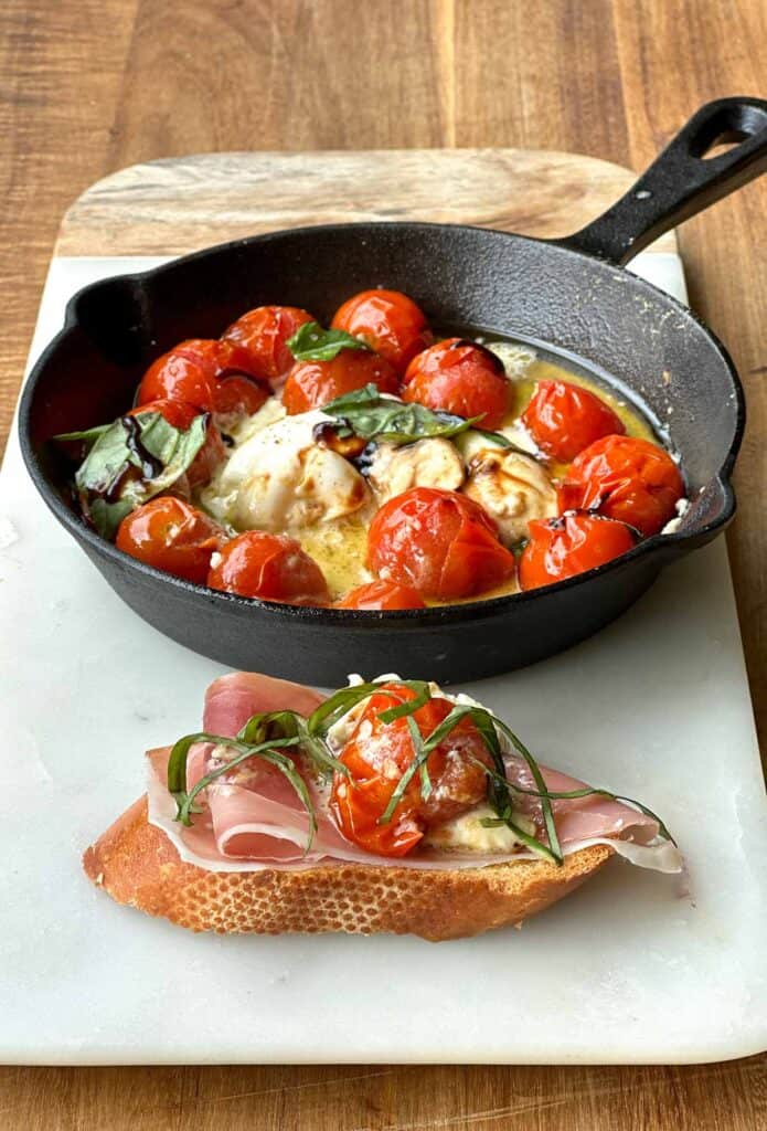 Toasted bread slice topped with prosciutto, roasted tomatoes, burrata cheese, and basil, cast iron skillet in the background.