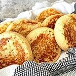 Golden brown wholemeal protein crumpets in a bowl with striped dish towel.