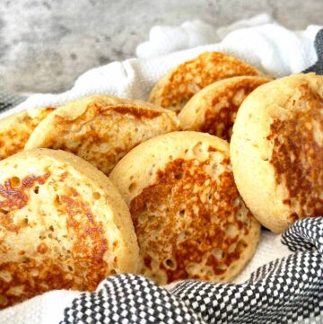 Golden brown wholemeal protein crumpets in a bowl with striped dish towel.