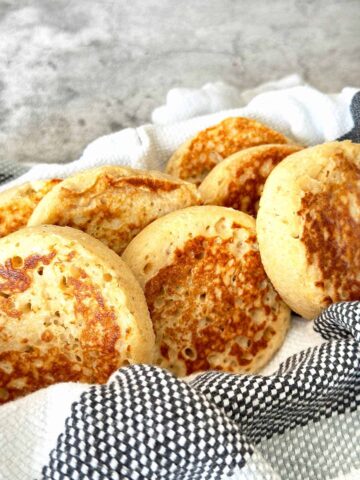 Golden brown wholemeal protein crumpets in a bowl with striped dish towel.