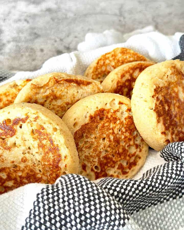 Golden brown wholemeal protein crumpets in a bowl with striped dish towel.