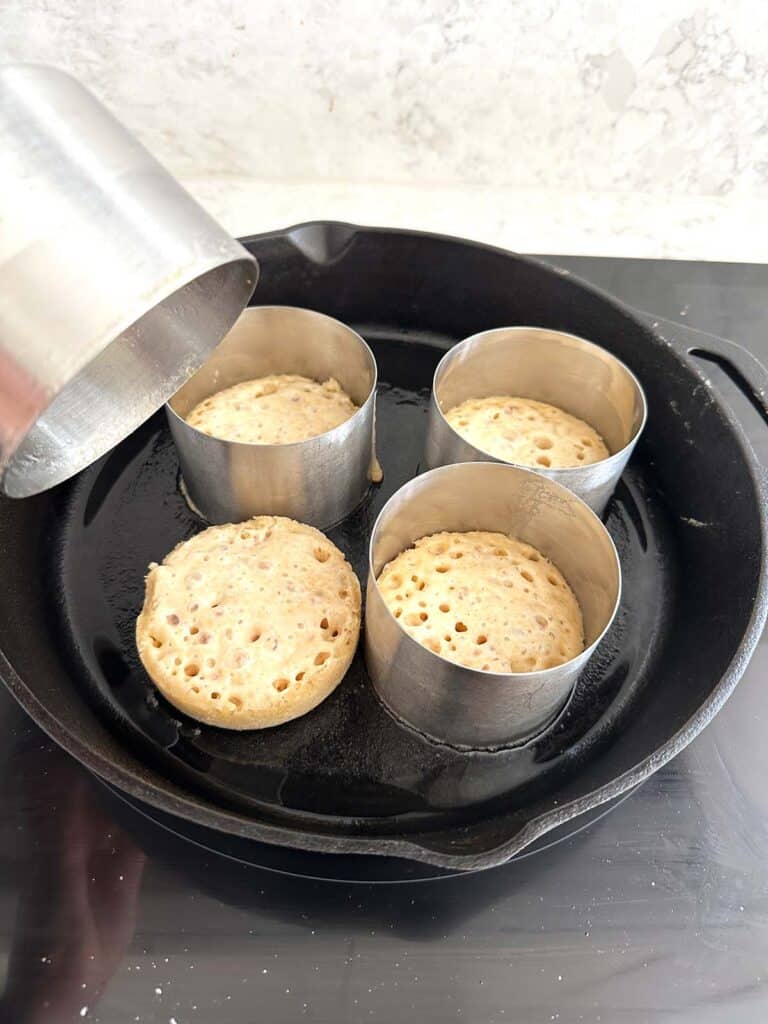 Four half-cooked crumpets in a black cast iron skillet, with three crumpets in metal rings and one ring removed.