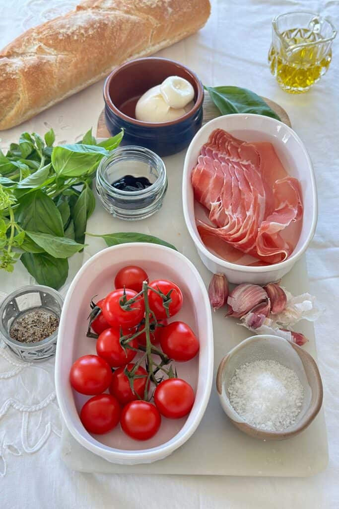 All the ingredients for burrata bruschetta: crusty bread slices, a ball of burrata cheese, halved cherry tomatoes, fresh basil leaves, a garlic clove, a small bowl of olive oil, and a pinch of salt and pepper.