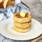 Stack of 3 fluffy crumpets topped with melting butter and honey on a white plate, crumpets, and honey spoon in background.