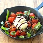 A cast iron skillet filled with roasted cherry tomatoes surrounding a ball of creamy burrata cheese, garnished with scattered fresh basil leaves and drizzled with balsamic glaze.