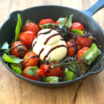 A cast iron skillet filled with roasted cherry tomatoes surrounding a ball of creamy burrata cheese, garnished with scattered fresh basil leaves and drizzled with balsamic glaze.