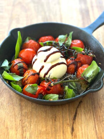 A cast iron skillet filled with roasted cherry tomatoes surrounding a ball of creamy burrata cheese, garnished with scattered fresh basil leaves and drizzled with balsamic glaze.