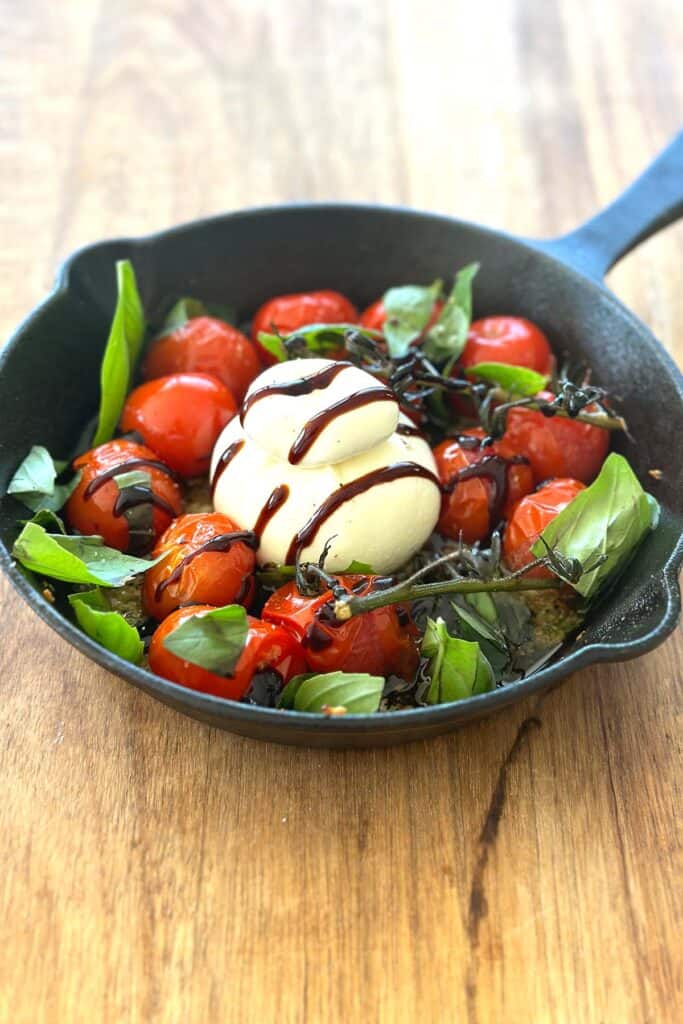 A cast iron skillet filled with roasted cherry tomatoes surrounding a ball of creamy burrata cheese, garnished with scattered fresh basil leaves and drizzled with balsamic glaze.