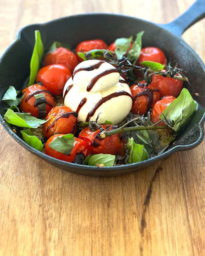 A cast iron skillet filled with roasted cherry tomatoes surrounding a ball of creamy burrata cheese, garnished with scattered fresh basil leaves and drizzled with balsamic glaze.