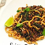 Beef udon noodles in a thick, flavourful sauce topped with fresh coriander, specks of chili, black sesame seeds, and a lime wedge on the side, on a white plate.