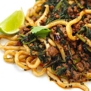 Beef udon noodles in a thick, flavourful sauce topped with fresh coriander, specks of chili, black sesame seeds, and a lime wedge on the side, on a white plate.