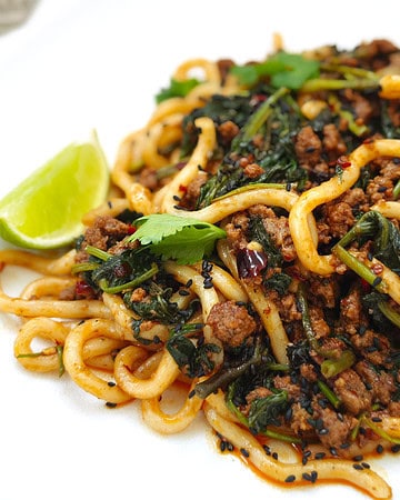 Beef udon noodles in a thick, flavourful sauce topped with fresh coriander, specks of chili, black sesame seeds, and a lime wedge on the side, on a white plate.