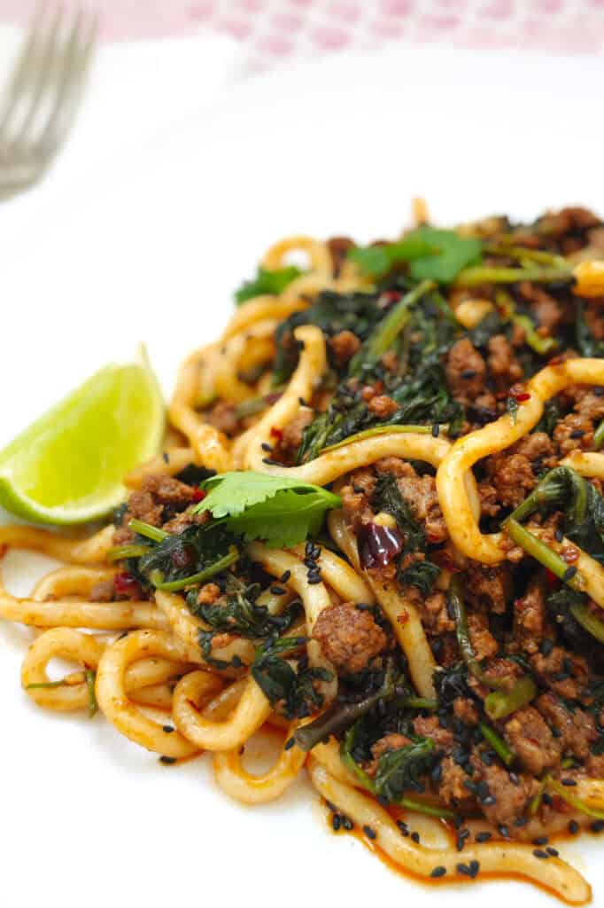 beef and noodles with ground beef in a thick, flavourful sauce topped with fresh coriander, specks of chili, black sesame seeds, and a lime wedge on the side, on a white plate.