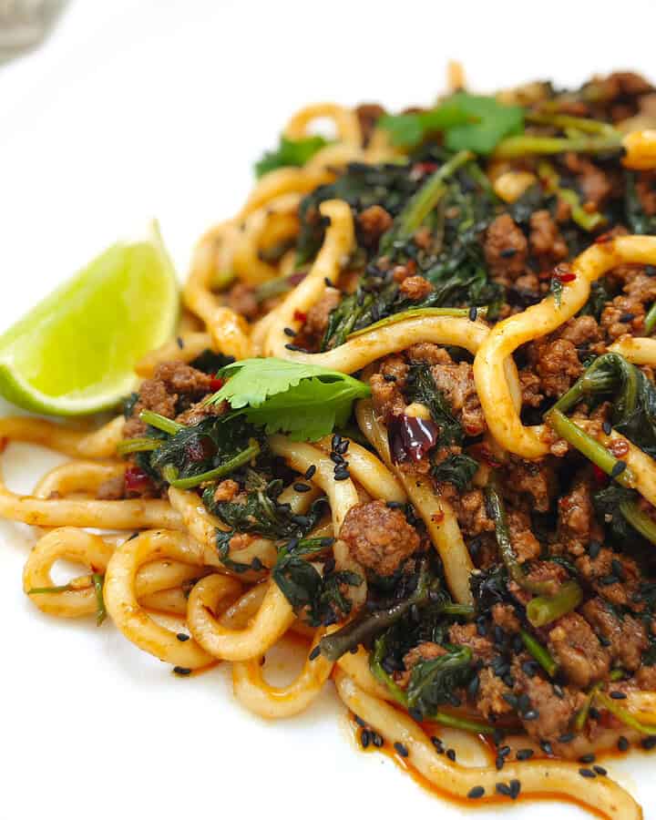 Beef udon noodles in a thick, flavourful sauce topped with fresh coriander, specks of chili, black sesame seeds, and a lime wedge on the side, on a white plate.