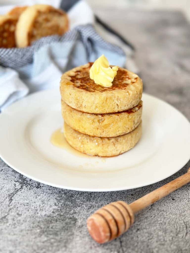 Stack of 3 fluffy crumpets topped with melting butter and honey on a white plate, crumpets, and honey spoon in background.