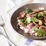 A brown plate with white rice topped with beef and onion stir fry, served with mushrooms, broccoli and coriander.