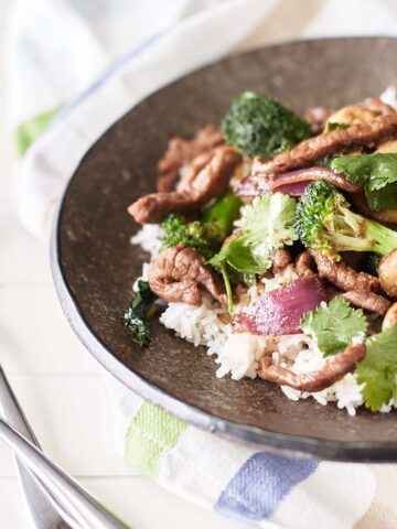 A brown plate with white rice topped with beef and onion stir fry, served with mushrooms, broccoli and coriander.