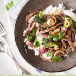 Stir-fried beef with broccoli, onions, and mushrooms on a grey plate, garnished with fresh coriander leaves.