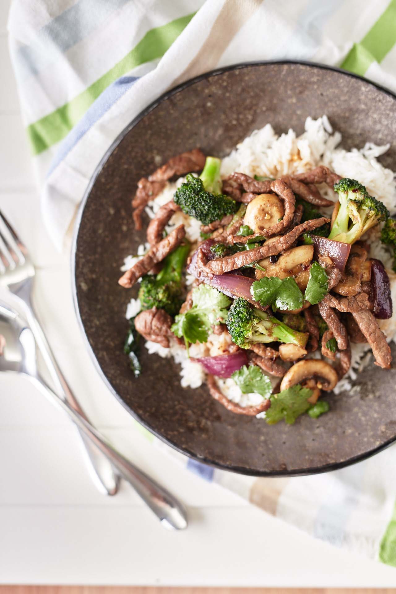 A brown plate with white rice topped with beef and onion stir fry, served with mushrooms, broccoli and coriander.