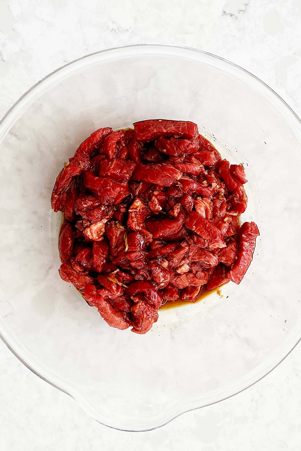 Glass bowl containing beef strips marinating in a dark, glossy sauce.