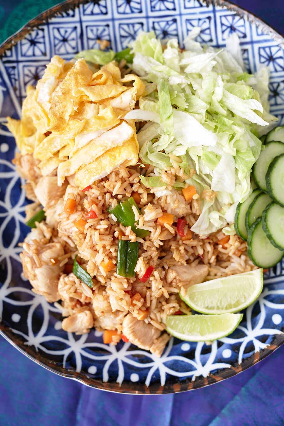 A plate of chicken nasi goreng with fried rice, sliced scrambled egg, fresh lettuce, cucumber, and lime wedges.