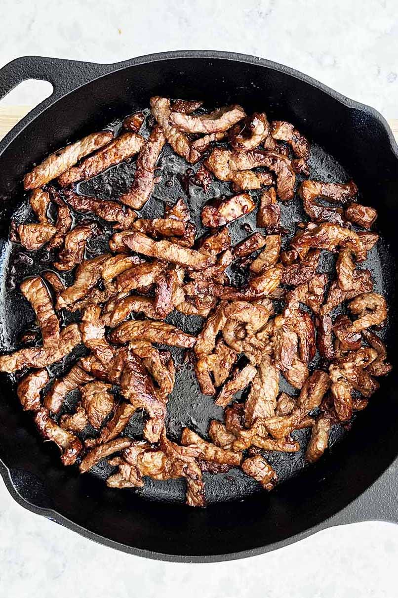 Caramelised beef strips in a cast iron skillet, glistening with a savoury sauce.