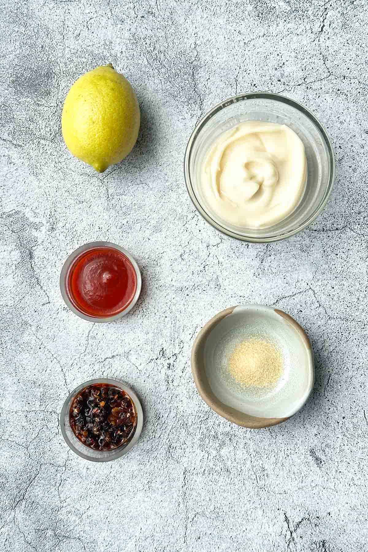 Five ingredients for Mayo chili (mayo, sriracha sauce, crispy chili oil, lemon, garlic powder) arranged on a bench.