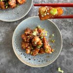 Two plates of crispy fried chicken bites coated in teriyaki sauce, garnished with sesame seeds and green onions, with one piece held by chopsticks, ready to be eaten.