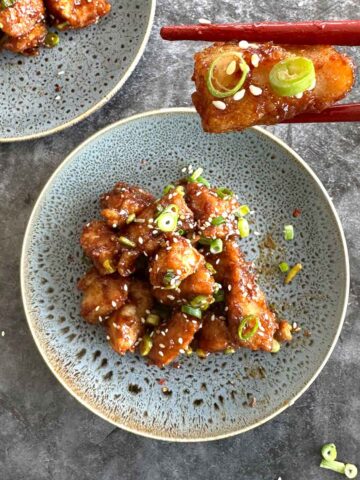 Two plates of crispy fried chicken bites coated in teriyaki sauce, garnished with sesame seeds and green onions, with one piece held by chopsticks, ready to be eaten.