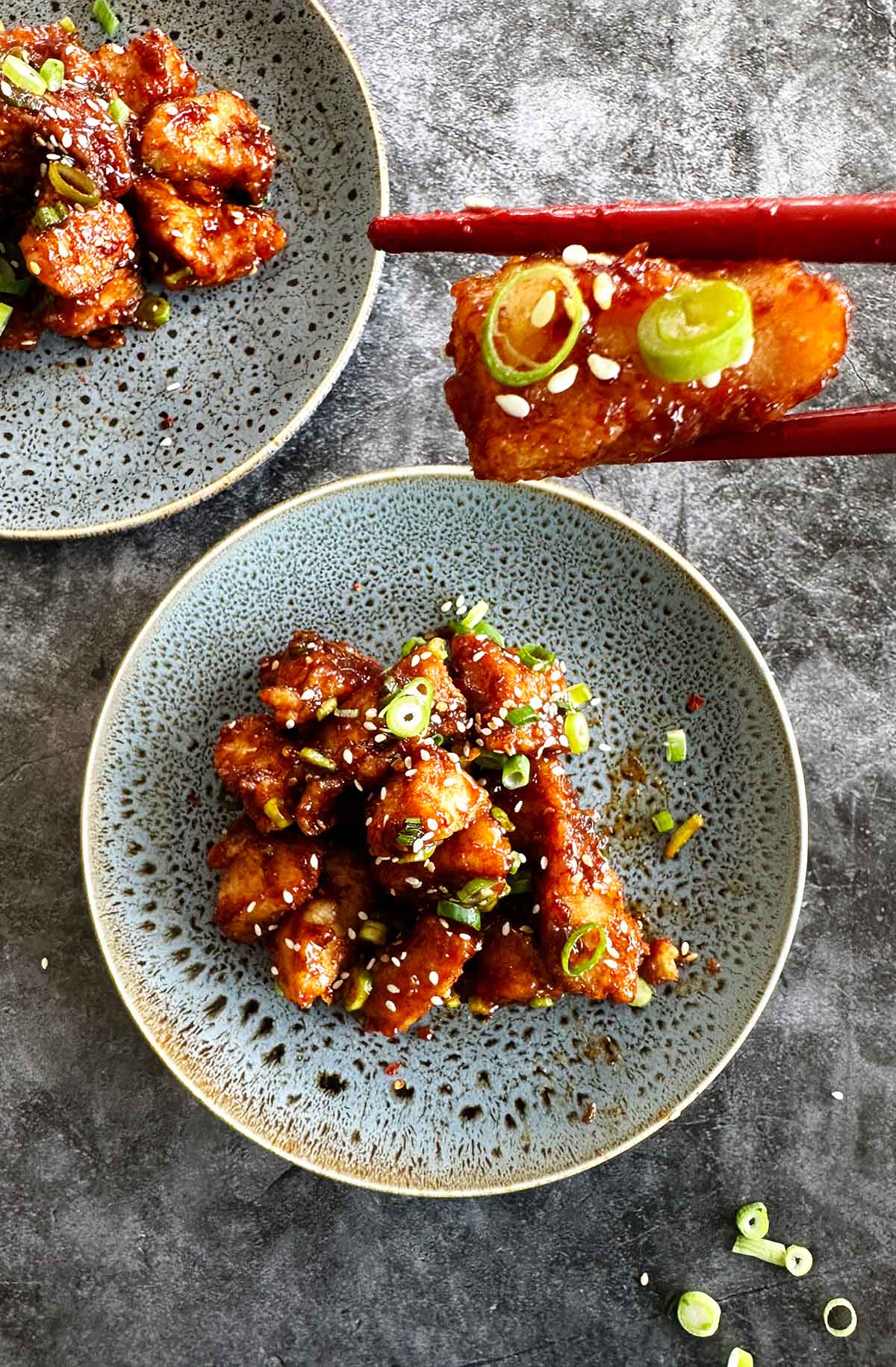 Two plates of crispy fried chicken bites coated in teriyaki sauce, garnished with sesame seeds and green onions, with one piece held by chopsticks, ready to be eaten.