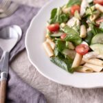 A colourful plate of chicken, pasta, spinach, cherry tomatoes, avocado, cucumber and fresh herbs