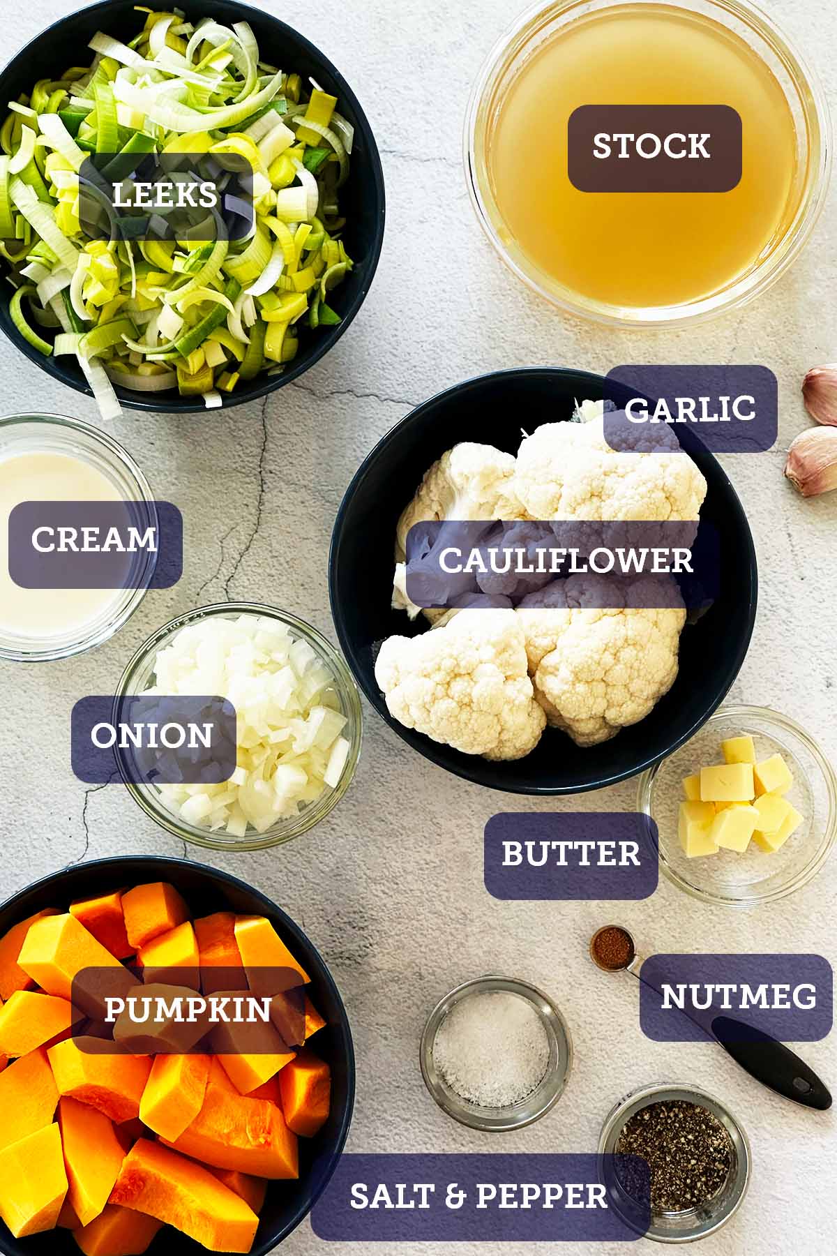 All ingredients for cauliflower and pumpkin soup portioned in small bowls, including chopped pumpkin, cauliflower, leek, onion, garlic, cream, stock, and garnishes.