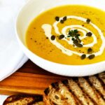 White bowl of cauliflower and pumpkin soup topped with Greek yoghurt and pepitas, served with toasted bread on the side.
