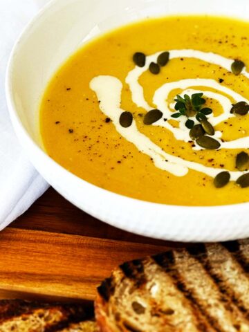White bowl of cauliflower and pumpkin soup topped with Greek yoghurt and pepitas, served with toasted bread on the side.
