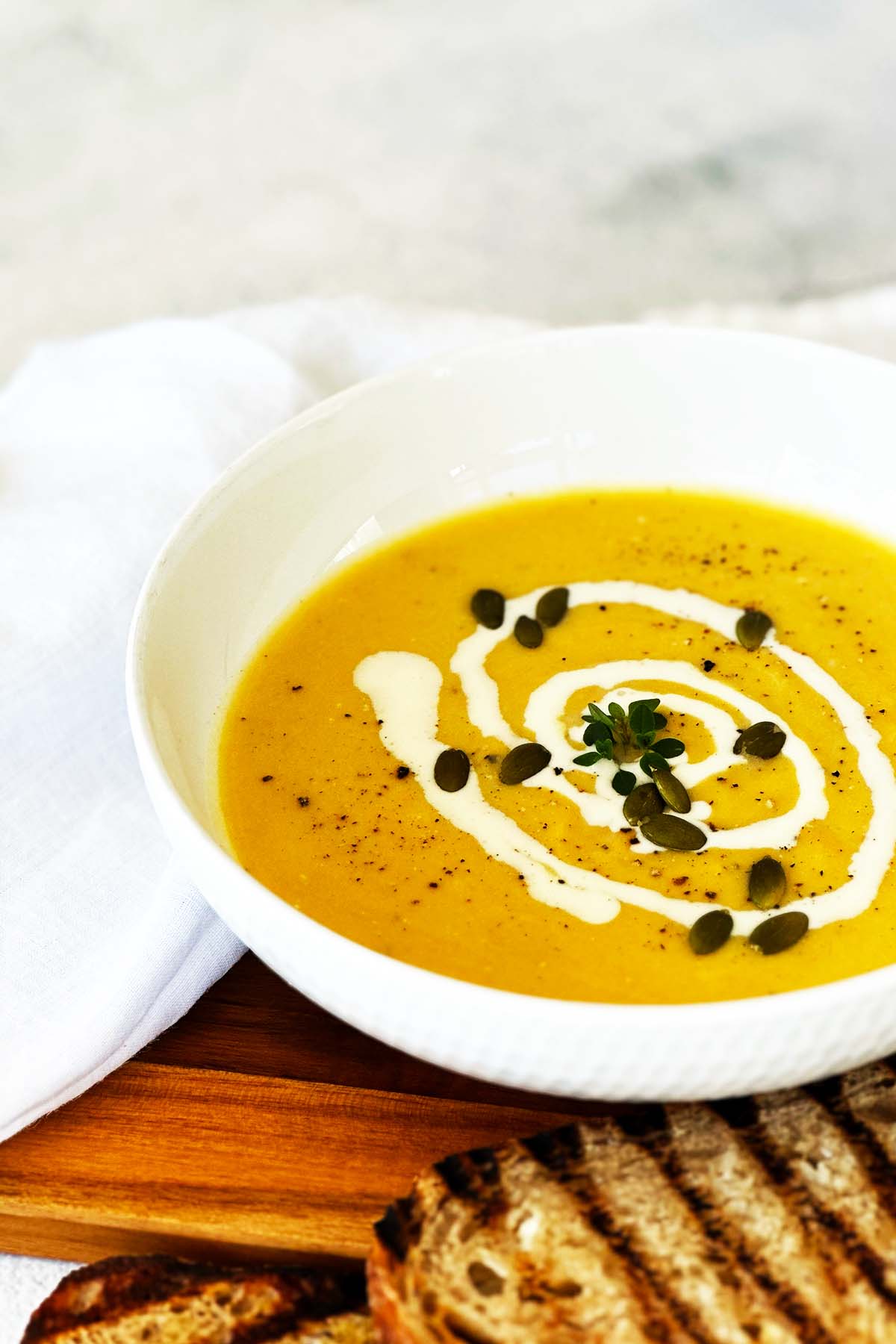 White bowl of cauliflower and pumpkin soup topped with Greek yoghurt and pepitas, served with toasted bread on the side.