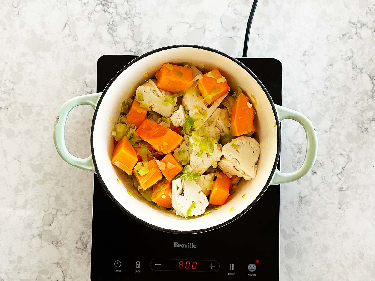 Chopped pumpkin and cauliflower added to sautéed aromatics in a pot as the next step in making soup.