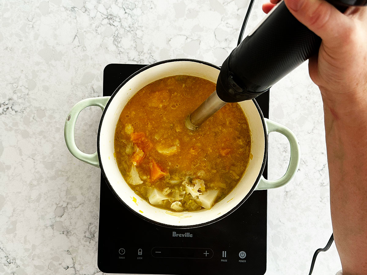 Blending cooked pumpkin and cauliflower soup with a stick blender until smooth and creamy.