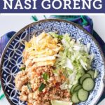 A plate of nasi goreng with chicken, fried rice, and sliced omelette, cucumber, lettuce and sliced lime on the side.