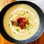 Creamy potato, leek and cauliflower soup served in a black bowl, garnished with candied bacon, swirls of cream, and fresh chives.