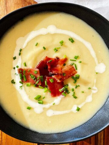 Creamy potato, leek and cauliflower soup served in a black bowl, garnished with candied bacon, swirls of cream, and fresh chives.