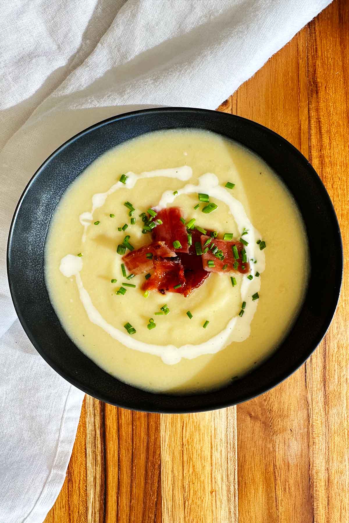 Creamy potato, leek and cauliflower soup served in a black bowl, garnished with candied bacon, swirls of cream, and fresh chives.