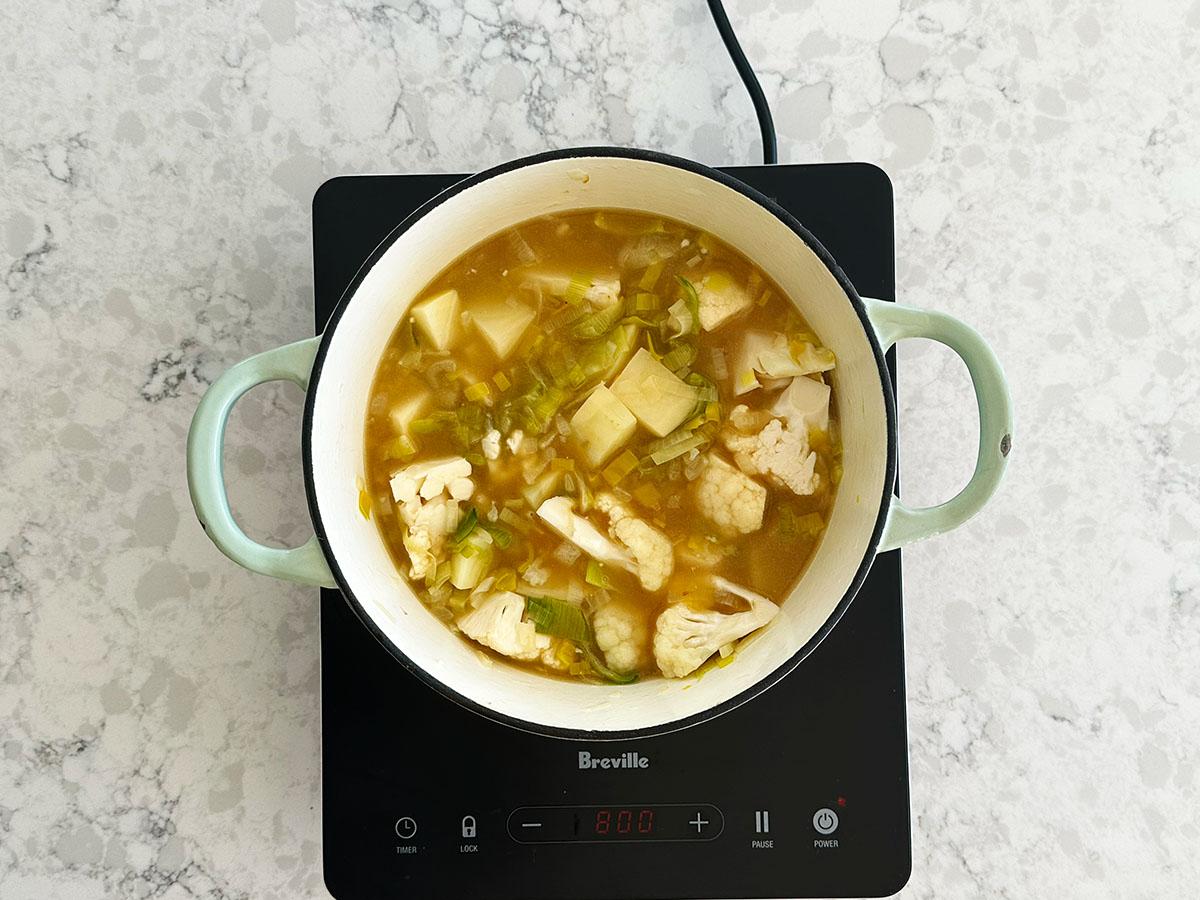 vegetable stock being poured into a pot with potatoes, cauliflower, leek, and onion to make the Vegetarian cauliflower potato soup