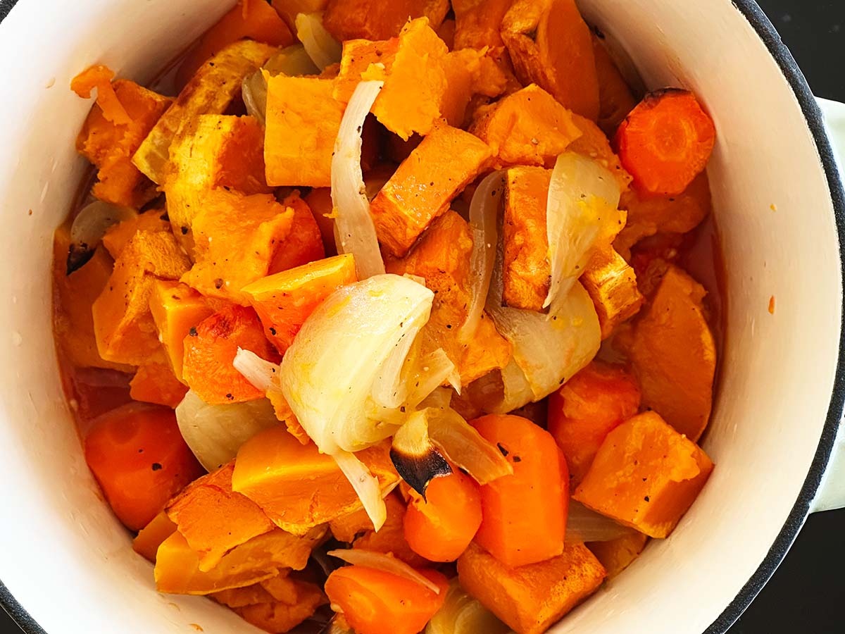Roasted butternut squash, tomatoes, carrots, and onion being added into a large pot for blending into soup.
