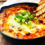 Baked chili cream cheese dip topped with green onions in a cast iron skillet, golden and bubbly, with a piece of crusty bread dipped into the warm, cheesy center.