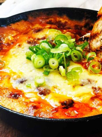 Baked chili cream cheese dip topped with green onions in a cast iron skillet, golden and bubbly, with a piece of crusty bread dipped into the warm, cheesy center.