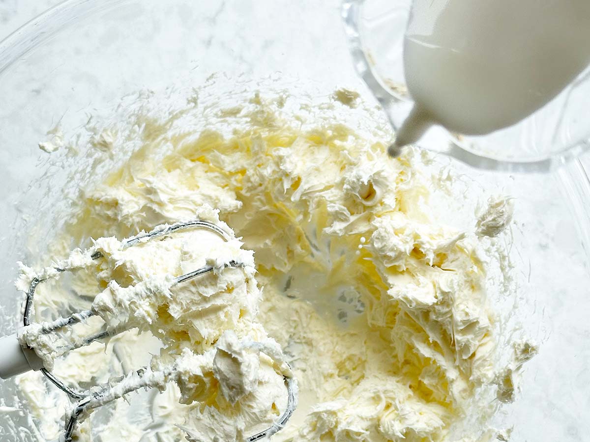 Adding cream or milk to whipped cream cheese in a glass bowl with a hand held electric mixer on the side.