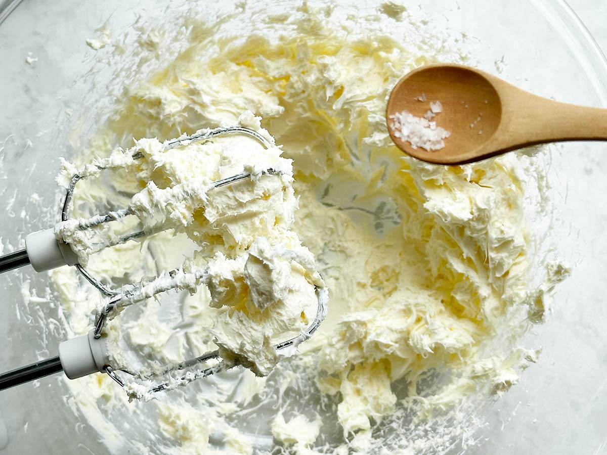 Adding salt to the cream cheese in a glass bowl with a hand held electric mixer on the side.