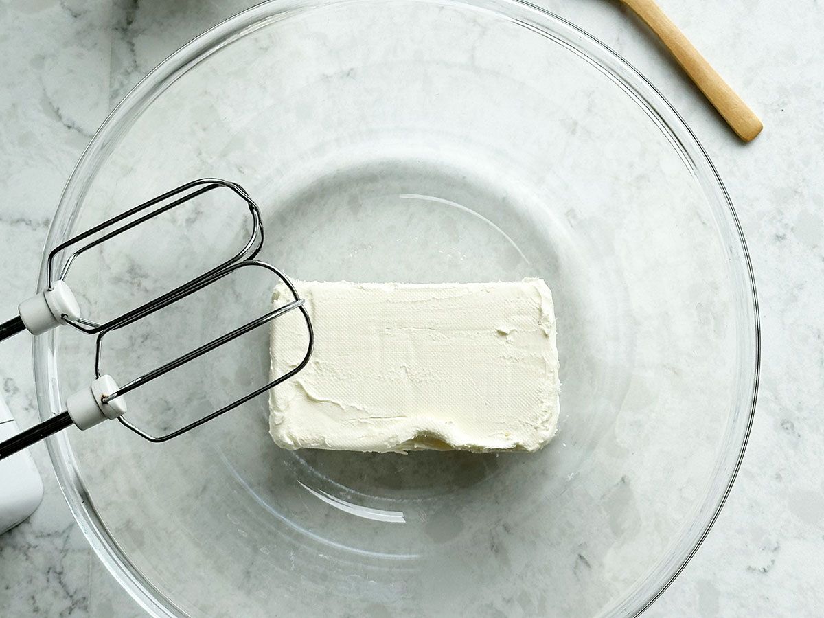Block of soften cream cheese in a glass bowl with a electric whisk on the side.
