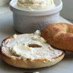 Whipped cream cheese spread on a toasted bagel with bowl in back ground and silver butter knife in front.