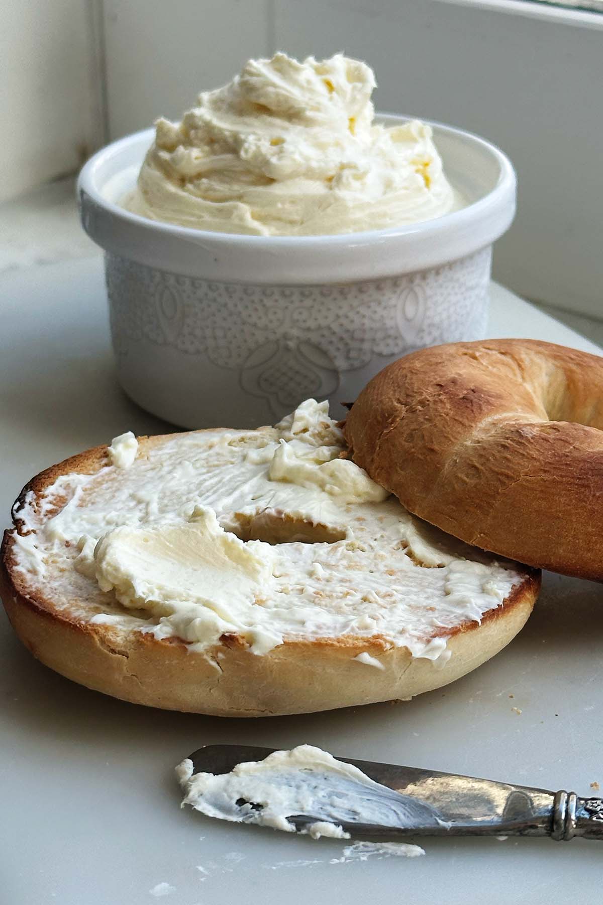 Cream cheese spread on a toasted bagel with bowl in back ground and silver butter knife in front.